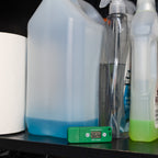 A HumidityPod in front of several cleaning supplies, including a large translucent jug with blue liquid, a clear spray bottle, and a white plastic bottle with a neon green liquid.