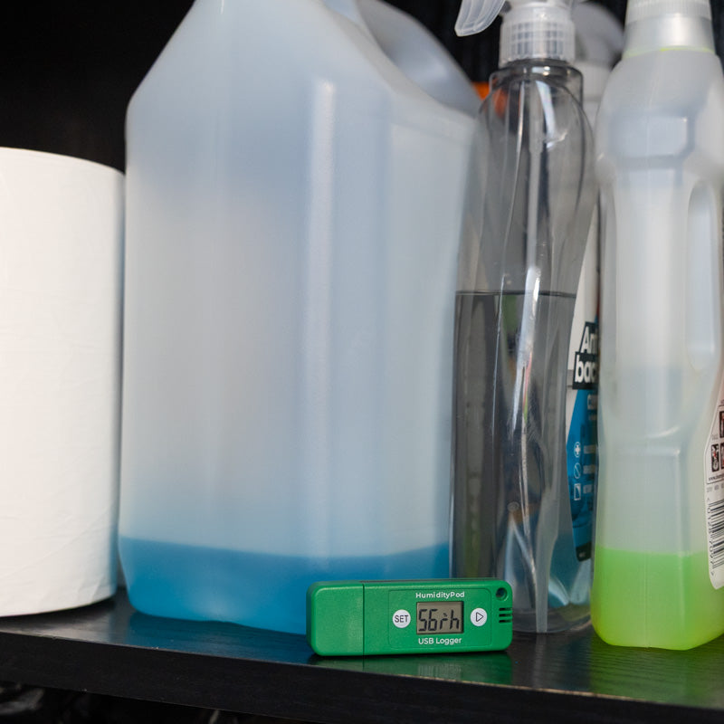 A HumidityPod in front of several cleaning supplies, including a large translucent jug with blue liquid, a clear spray bottle, and a white plastic bottle with a neon green liquid.