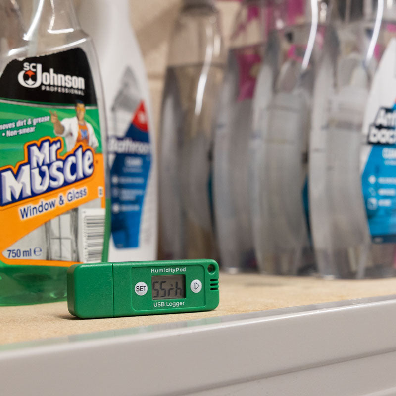 A HumidityPod on a shelf with household cleaning products, including a prominent bottle of Window & Glass cleaner.