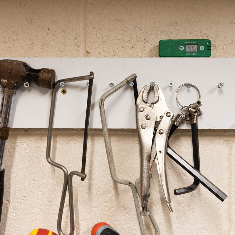 A set of hand tools including a hacksaw, pliers, and a clamp hook mounted on a white horizontal board against a beige textured wall with a HumidityPod