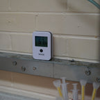 A white digital temperature and humidity NODE model on a a metal shelf against a beige brick wall in a manufacturing setting