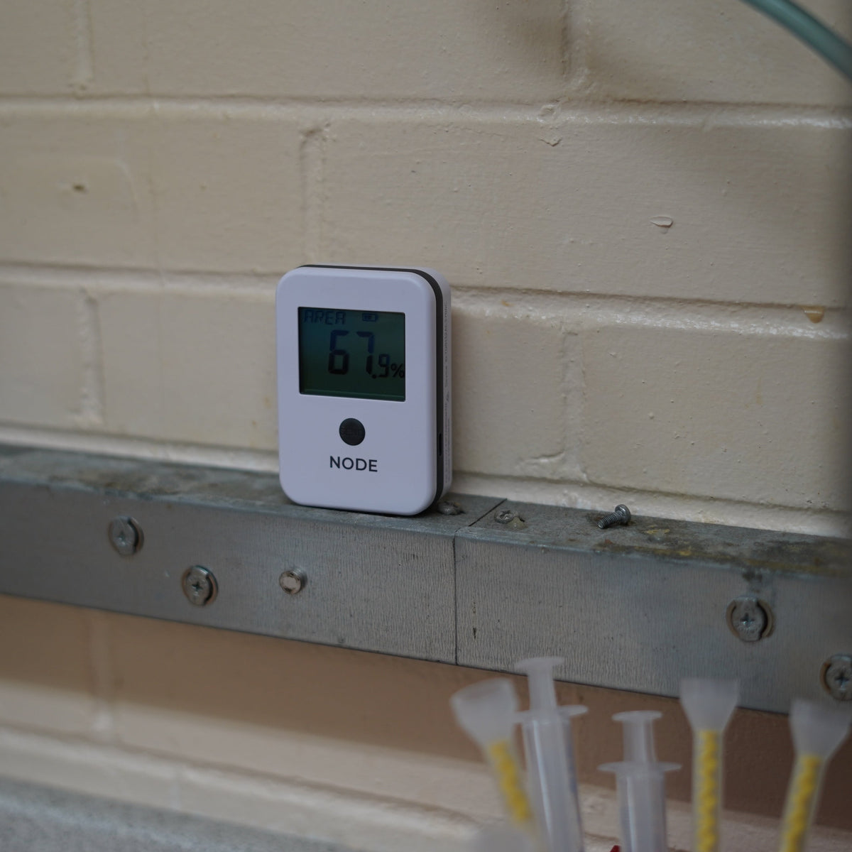 A white digital temperature and humidity NODE model on a a metal shelf against a beige brick wall in a manufacturing setting