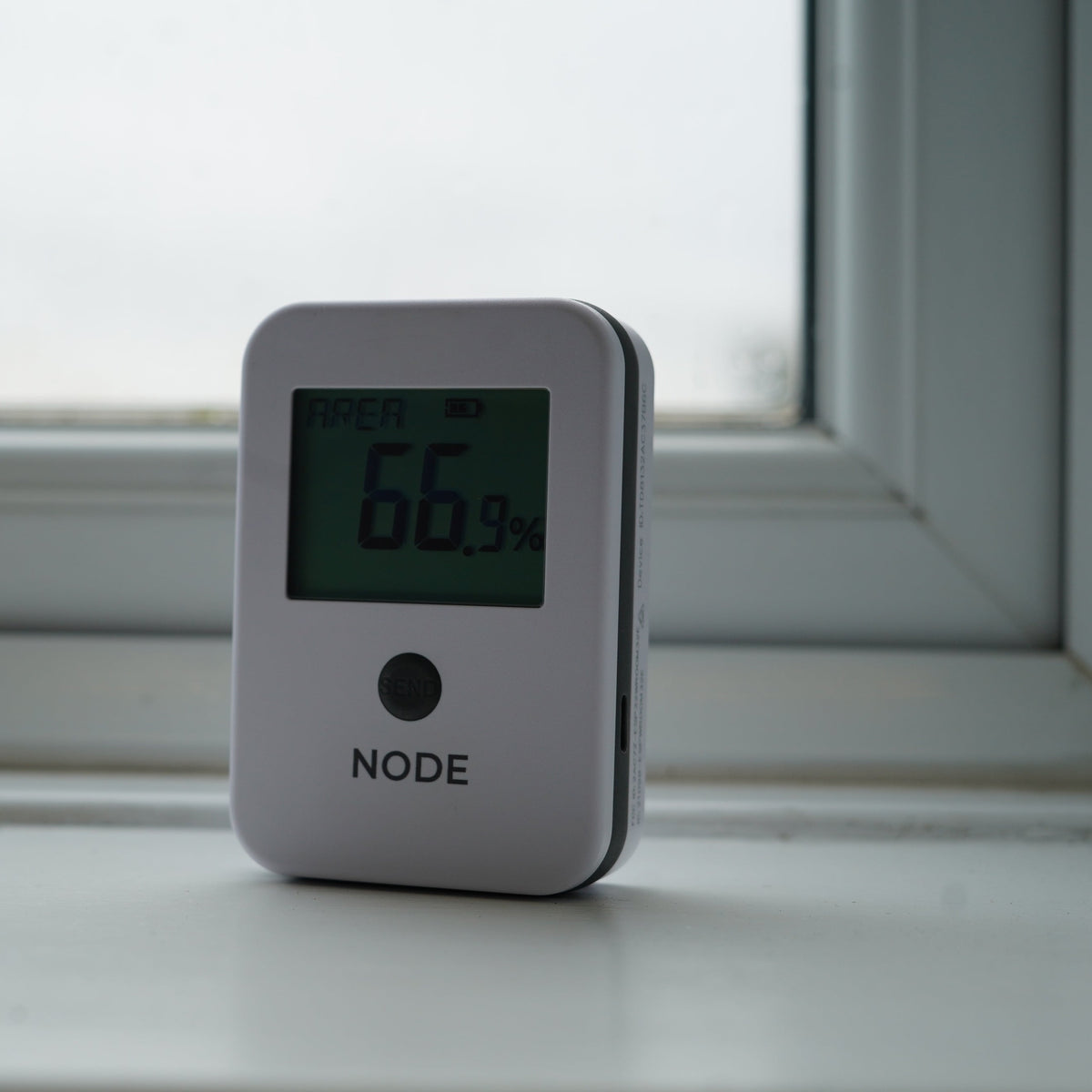 A white NODE humidity and temperature sensor with a digital display showing 66% humidity, sitting on a white windowsill in front of a window