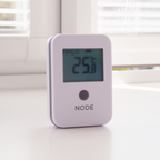 A small white electronic device labeled NODE with a digital display reading 25°C, standing on a white tabletop near a window in an office.