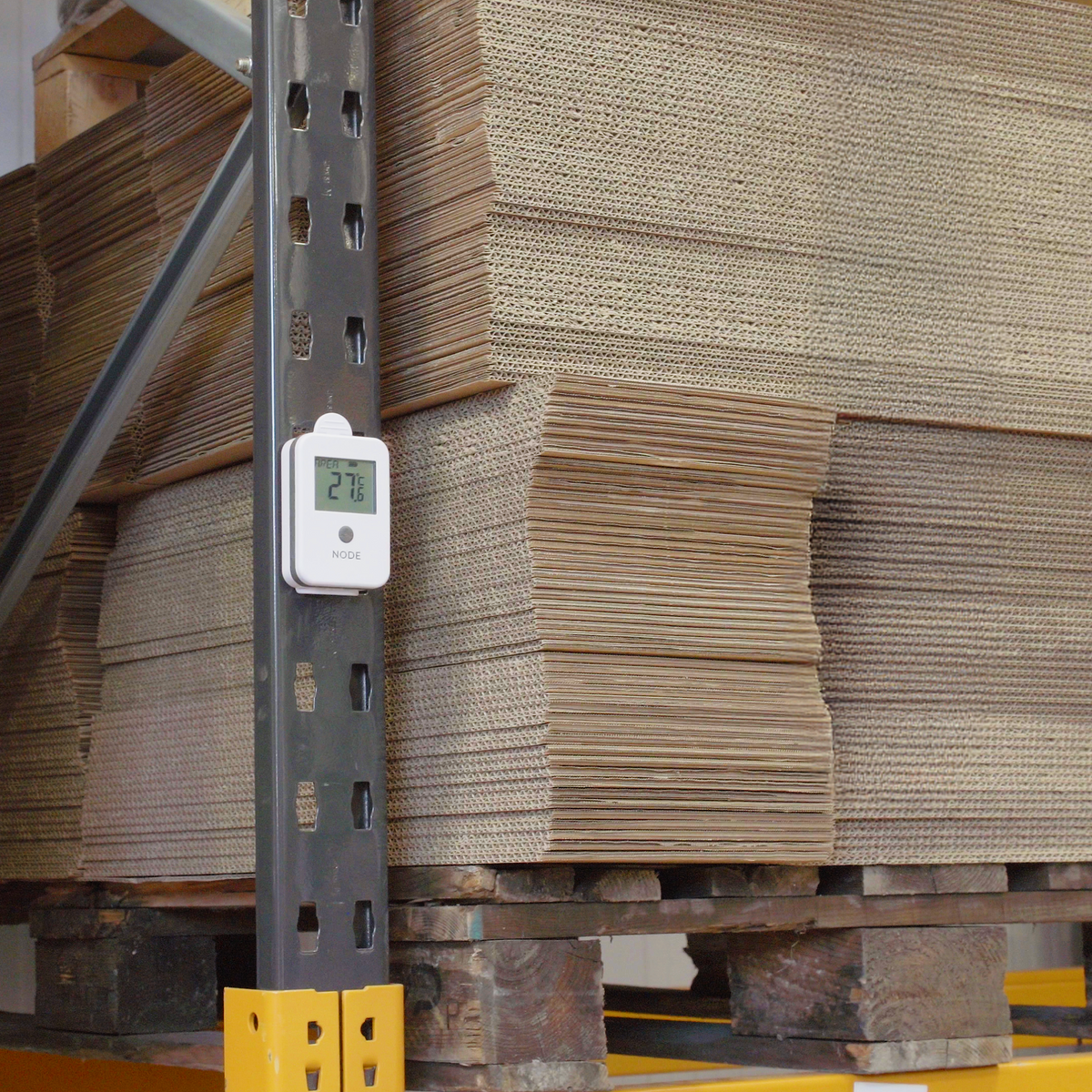A white digital temperature sensor mounted on a black metal warehouse rack, with stacks of corrugated cardboard boxes on wooden pallets in the background.