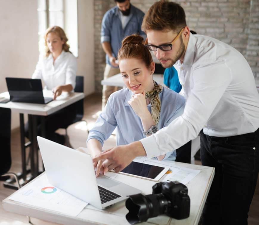 Two people working on a laptop with another person in the background.