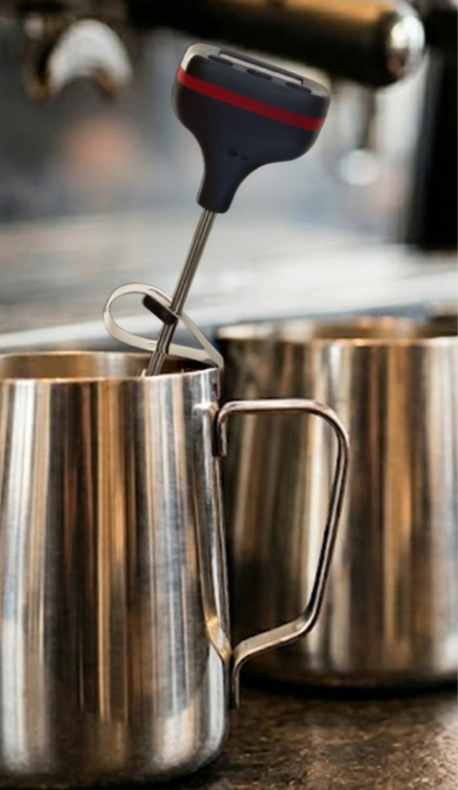 Foam mixer being used in a metal pitcher with a blurred kitchen background and a milk thermometer