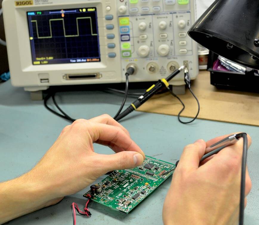 Person working on a circuit board with an oscilloscope in the background
