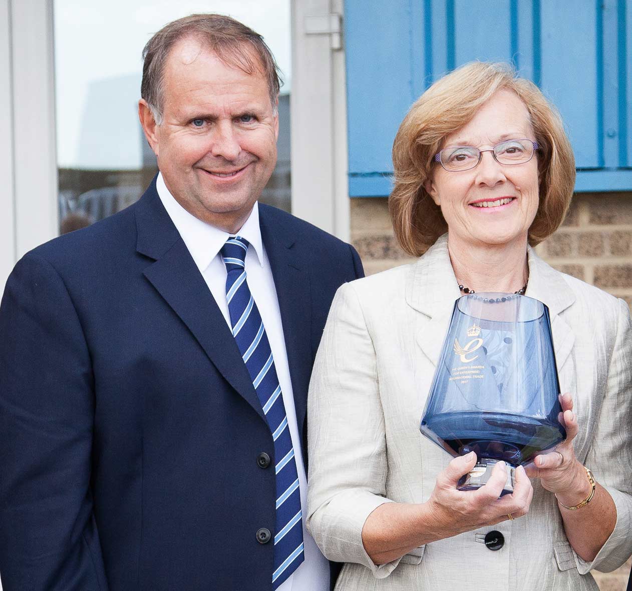 Peter Webb in a suit and tie smiling next to Miriam Webb wearing glasses, beige blazer, smiling and holding the Queen's award 