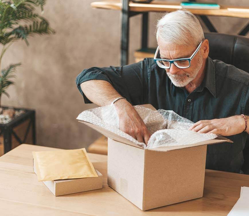 Gray haired man with glasses looking into a cardboard box with bubblewrap in it and some packages on the side of the box he is looking in