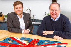 Jason and Peter Webb sitting behind a table smiling with Thermapens layed in UK flag pattern infront of them